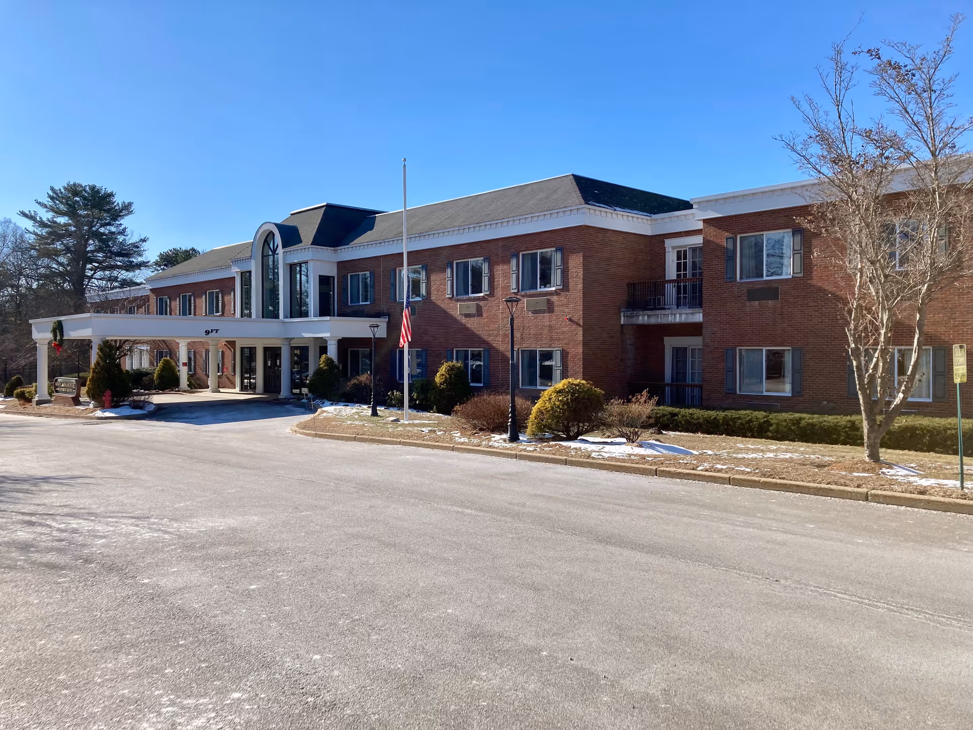 Exterior view of a two-story brick building with multiple windows and a covered entrance. There is a flagpole with an American flag in front, some small bushes and trees around the building, and a clear blue sky above.