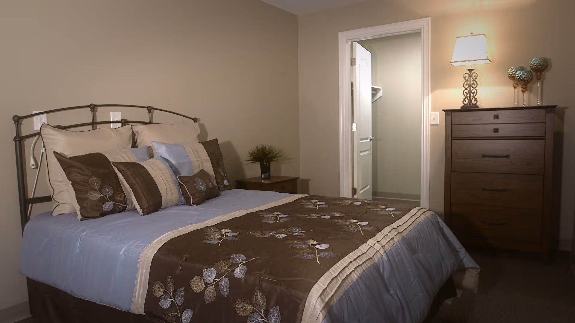 A neatly made bedroom with a bed, multiple pillows, a nightstand, a dresser with a lamp, and an open closet door.