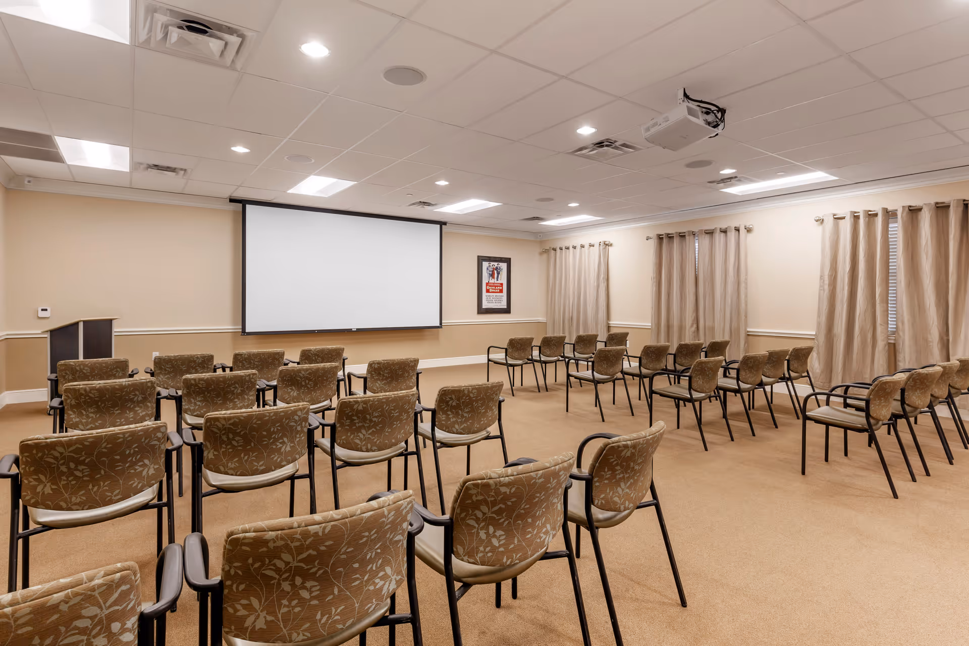 A conference or meeting room with rows of patterned chairs facing a large white projection screen mounted on a beige wall. The room has beige carpeting, beige walls with white trim, and several windows covered with beige curtains. A podium is positioned to the left side of the screen, and a ceiling-mounted projector is visible.