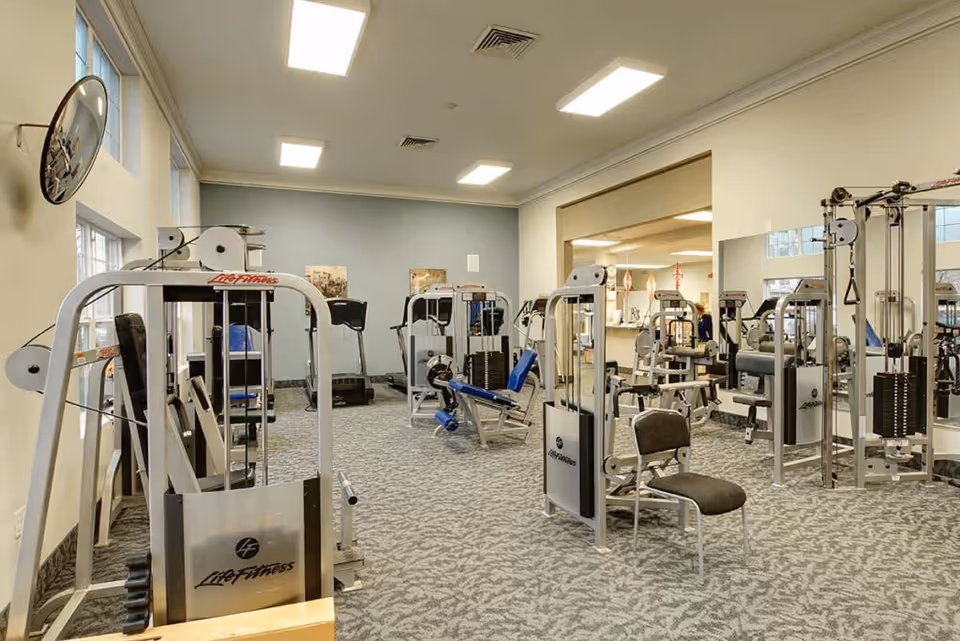 A well-equipped exercise room with weight machines, cardio equipment, and mirrors in a senior living facility.