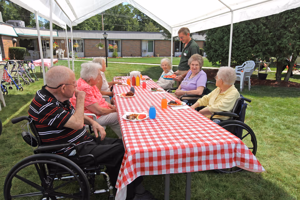 A group of elderly people, some in wheelchairs, sitting around a long table covered with a red and white checkered tablecloth under a white canopy outdoors. They are enjoying a meal with plates of food and colorful drinks. A man is serving watermelon slices to the group. The setting is a grassy courtyard with a brick building and trees in the background.