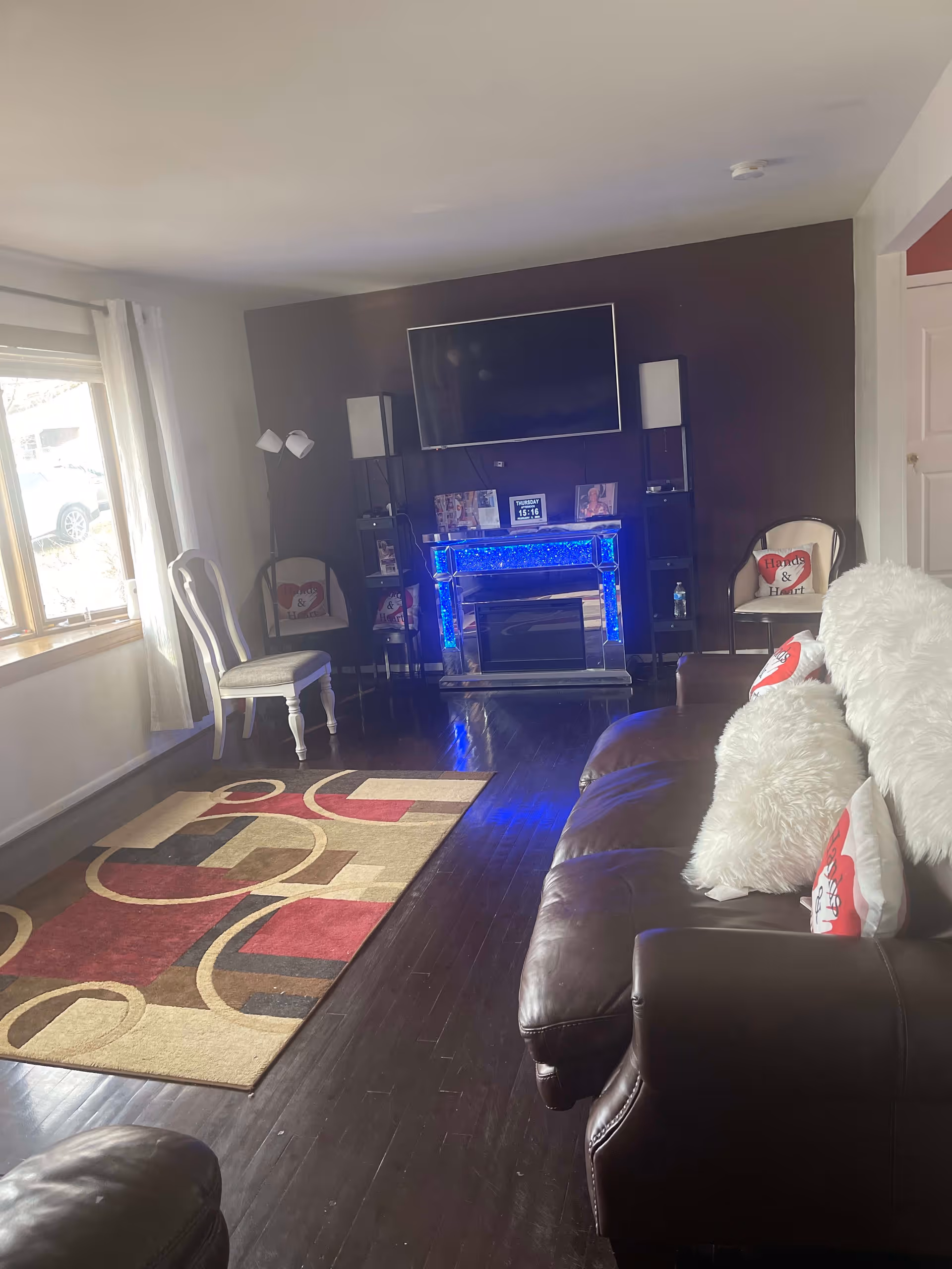 A cozy living room with dark hardwood floors, a patterned area rug, a brown leather couch with white fluffy pillows, and a wall-mounted flat screen TV above a blue-lit electric fireplace. There are two chairs with heart-themed cushions and a floor lamp near a window with white curtains letting in natural light.