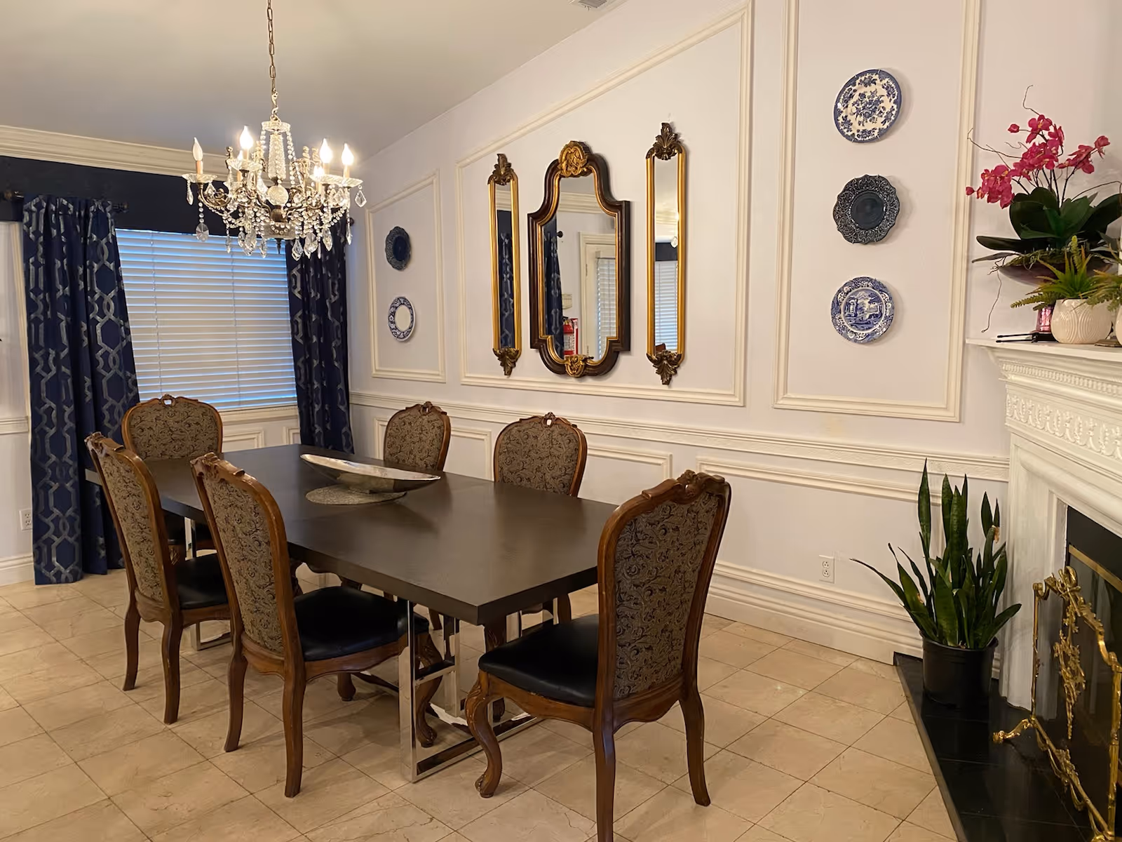 Formal dining room with a dark rectangular table surrounded by ornate chairs, a crystal chandelier, mirrors and decorative plates on paneled walls.