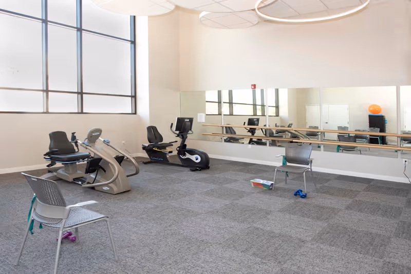 Bright fitness room with recumbent exercise bikes, chairs, a large wall mirror and small workout equipment on a carpeted floor.