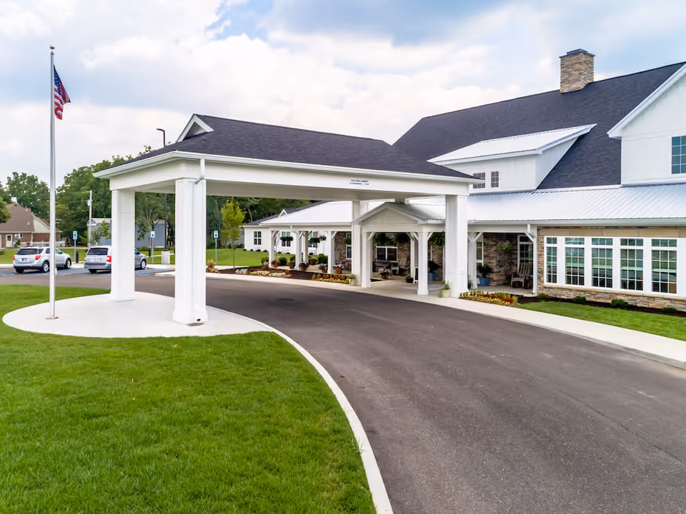 Entrance driveway of a senior living facility with a covered drop-off area, an American flag on a flagpole, and a large building with white siding, stone accents, and multiple windows under a partly cloudy sky.