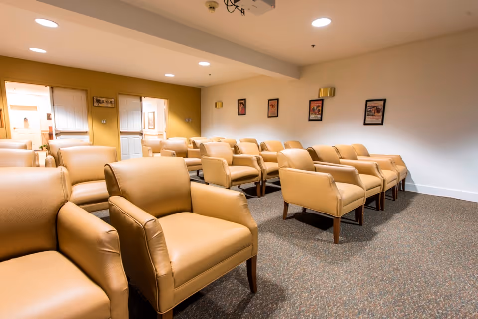 A room with multiple tan leather armchairs arranged in rows facing the front, beige walls with framed pictures, and a ceiling-mounted projector, suggesting a meeting or presentation space.