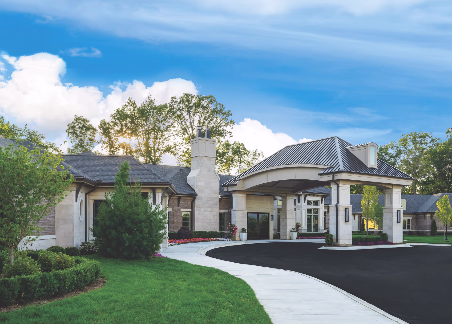 Exterior view of a senior living facility building with a covered entrance, surrounded by green grass, trees, and landscaping under a blue sky with clouds.