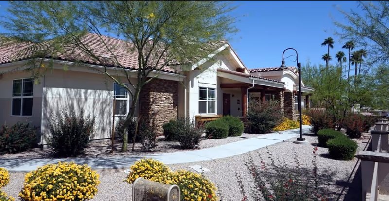 Exterior view of a single-story building with beige walls and a red tile roof, surrounded by desert landscaping including small bushes, yellow flowers, and trees under a clear blue sky.