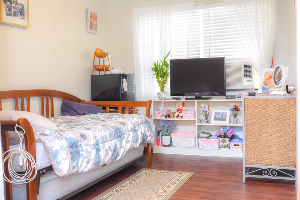 A cozy bedroom with a wooden daybed covered in a floral comforter and pillows. Next to the bed is a small black refrigerator with a basket on top. Across from the bed is a white shelving unit holding a flat-screen TV, an air conditioner, framed photos, decorative plants, and various storage baskets. A wicker cabinet with a clock and other small items is positioned to the right. The room has wooden flooring and a window with white blinds and sheer curtains letting in natural light.