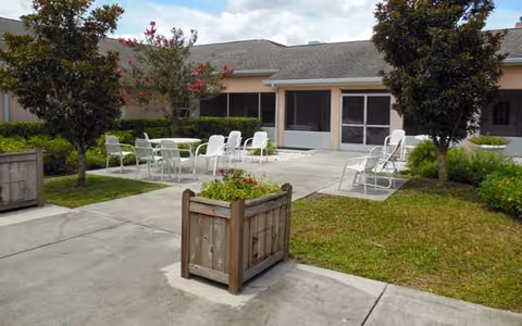 Outdoor patio area with white metal chairs and tables arranged on a concrete surface surrounded by grass and trees, adjacent to a building with screened windows and doors.
