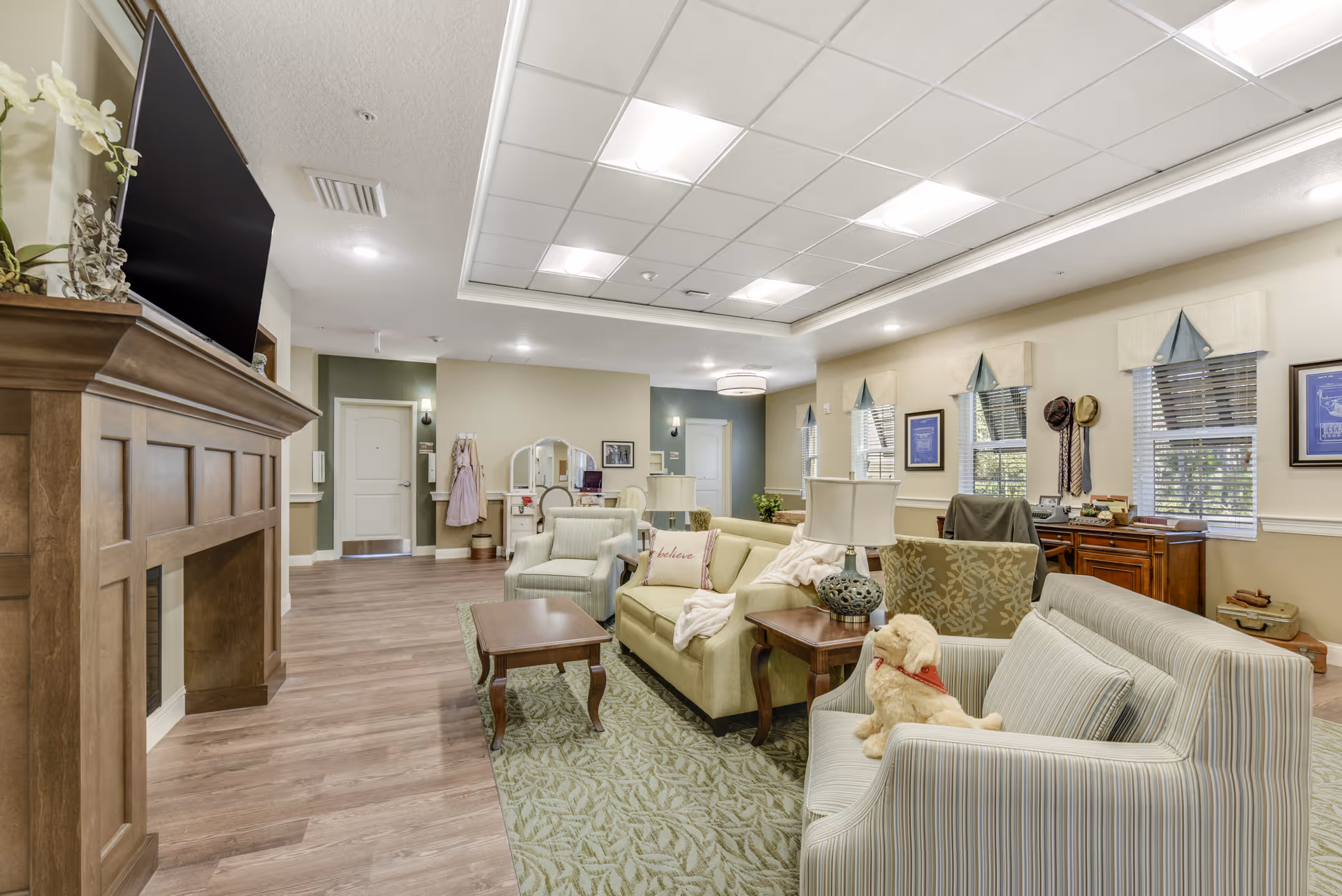 A spacious senior living facility common area with comfortable seating including armchairs and sofas arranged around wooden coffee tables. The room features a wooden fireplace with a large flat-screen TV mounted above it. The walls are painted in soft neutral tones with several windows letting in natural light. There are decorative elements such as framed pictures, a vanity table with a mirror, and a small stuffed dog on one of the sofas. The flooring is a combination of wood and a patterned area rug.