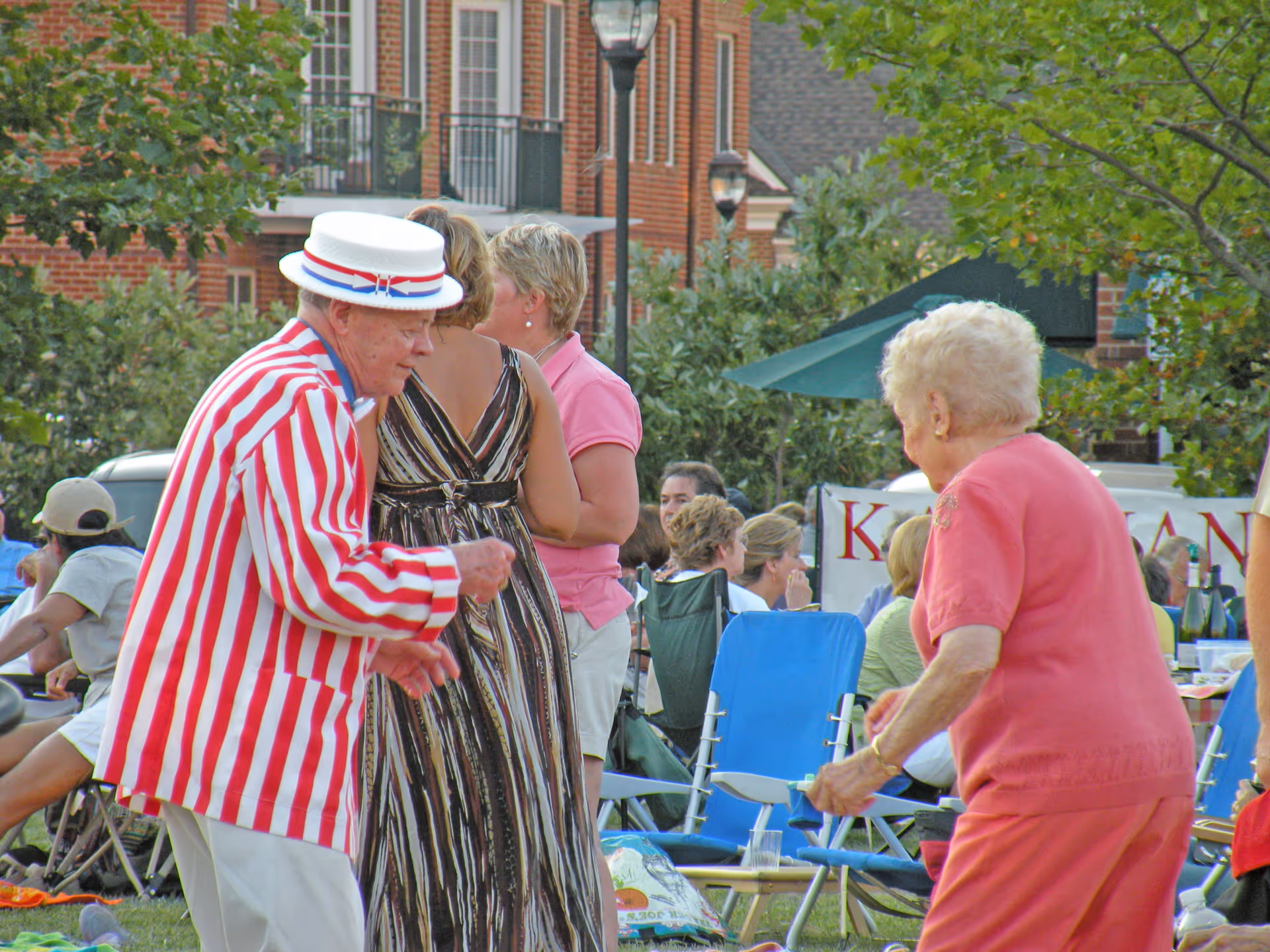An elderly man in a red and white striped jacket and white hat dances outdoors with an elderly woman in a pink outfit. In the background, several people are seated on lawn chairs near a brick building and trees.