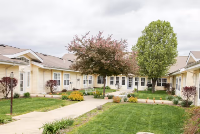 Courtyard with a paved walkway, green lawn, flowering trees, benches, and surrounding single-story yellow buildings.