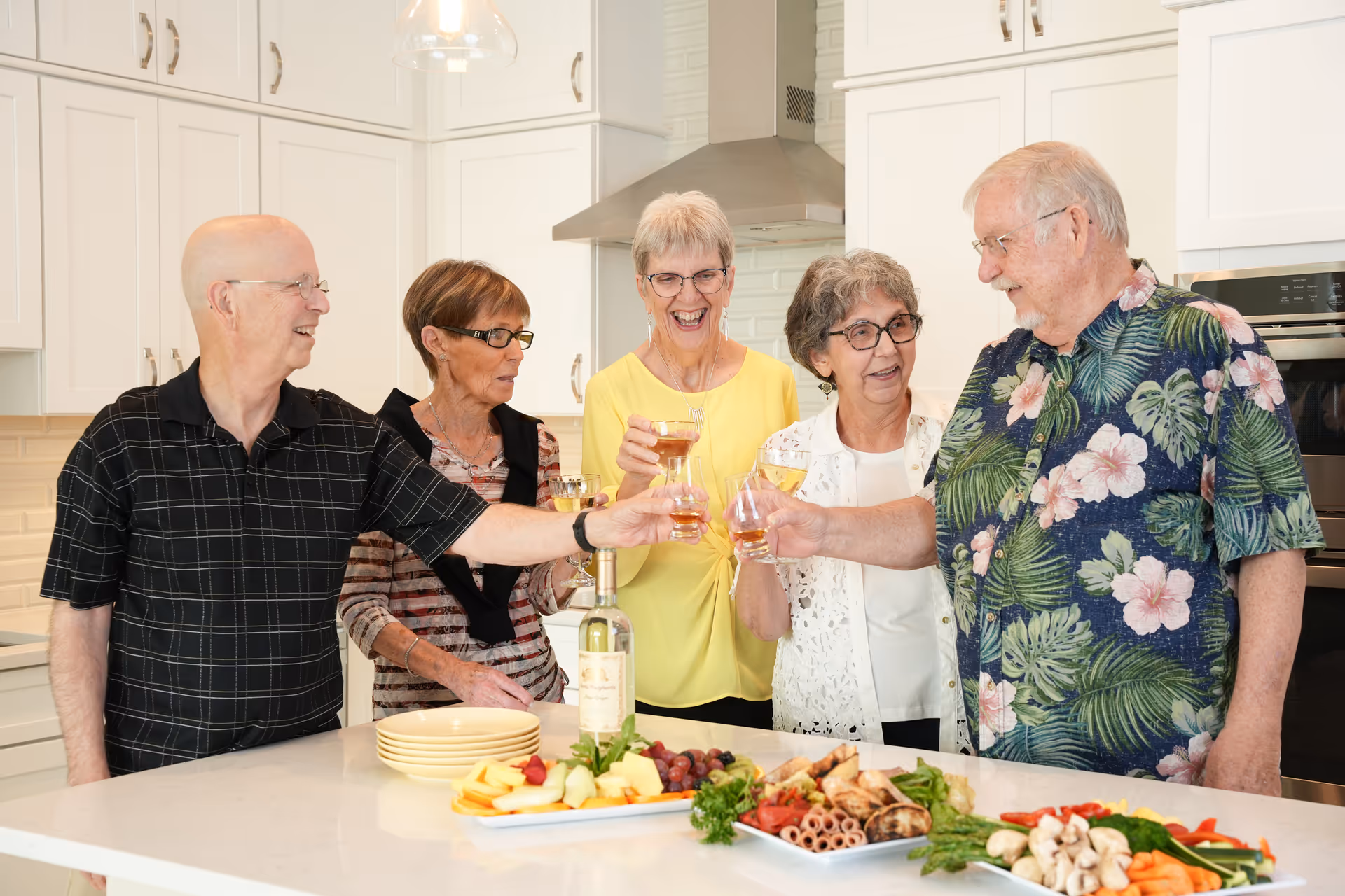 Five elderly people standing around a kitchen island, smiling and toasting with glasses of wine. The kitchen has white cabinets and a modern stainless steel range hood. On the counter, there are plates and trays with assorted fruits, vegetables, and appetizers.