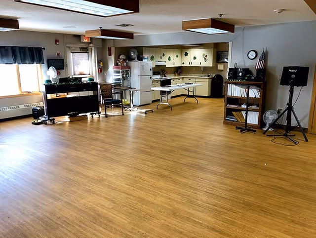 A spacious room with wooden flooring featuring a kitchen area in the background with white cabinets decorated with green shamrocks. The kitchen includes a refrigerator, stove, and a folding table. There are chairs, a piano, a bookshelf with books and a clock, and various fans placed around the room. The room is well-lit with natural light coming from a window with a dark valance.