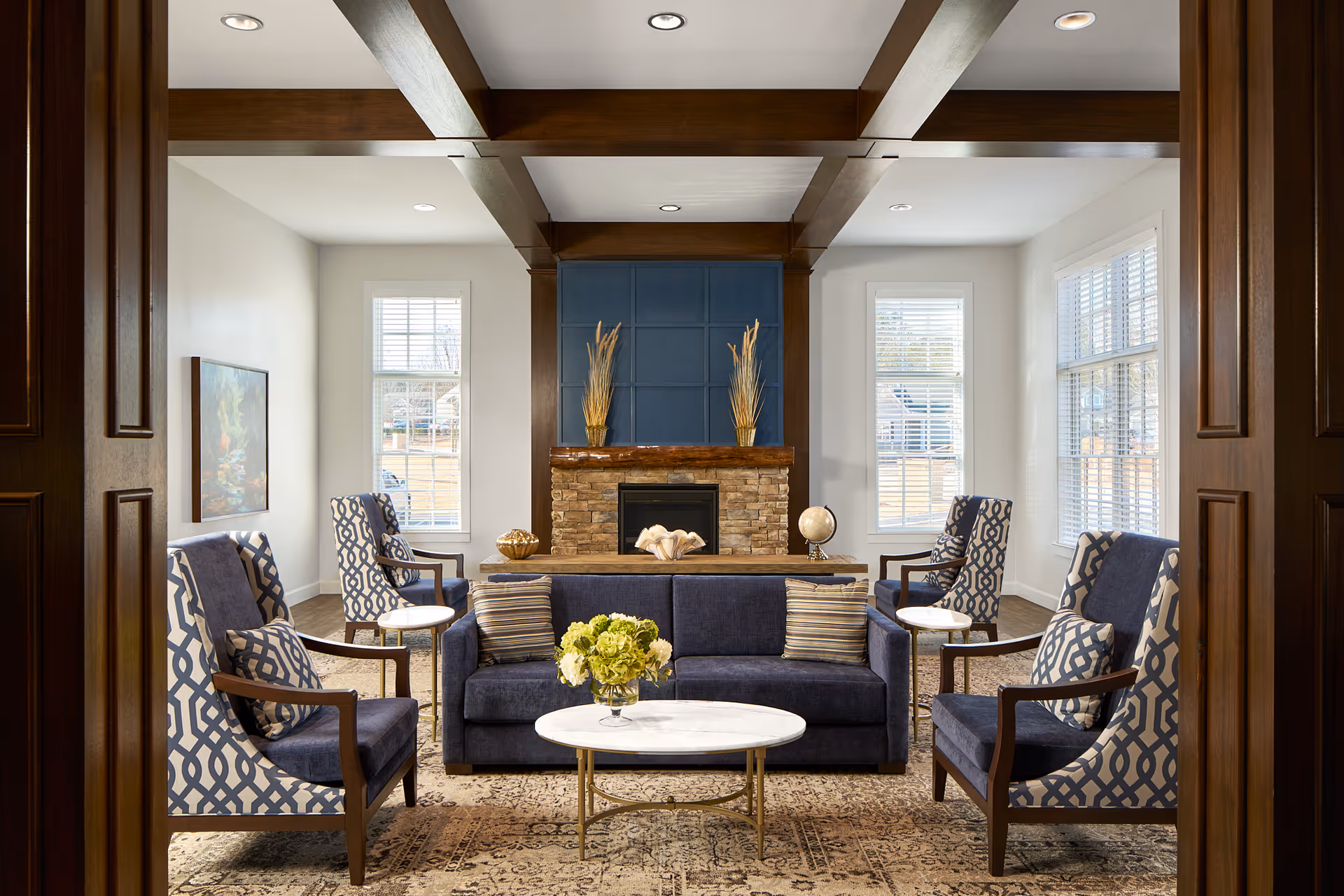 A cozy living room with dark blue upholstered chairs and a matching sofa arranged around a white marble coffee table. The room features a stone fireplace with a wooden mantel and decorative vases, flanked by two windows with white blinds. The ceiling has wooden beams, and the floor is covered with a patterned rug.
