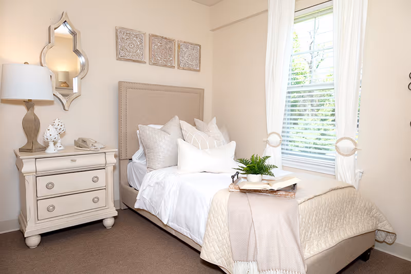 A cozy bedroom with a beige upholstered bed adorned with multiple pillows and a beige quilt. A small wooden nightstand with a lamp, decorative items, and a telephone is beside the bed. A window with white curtains lets in natural light, and three decorative wall hangings are above the bed.