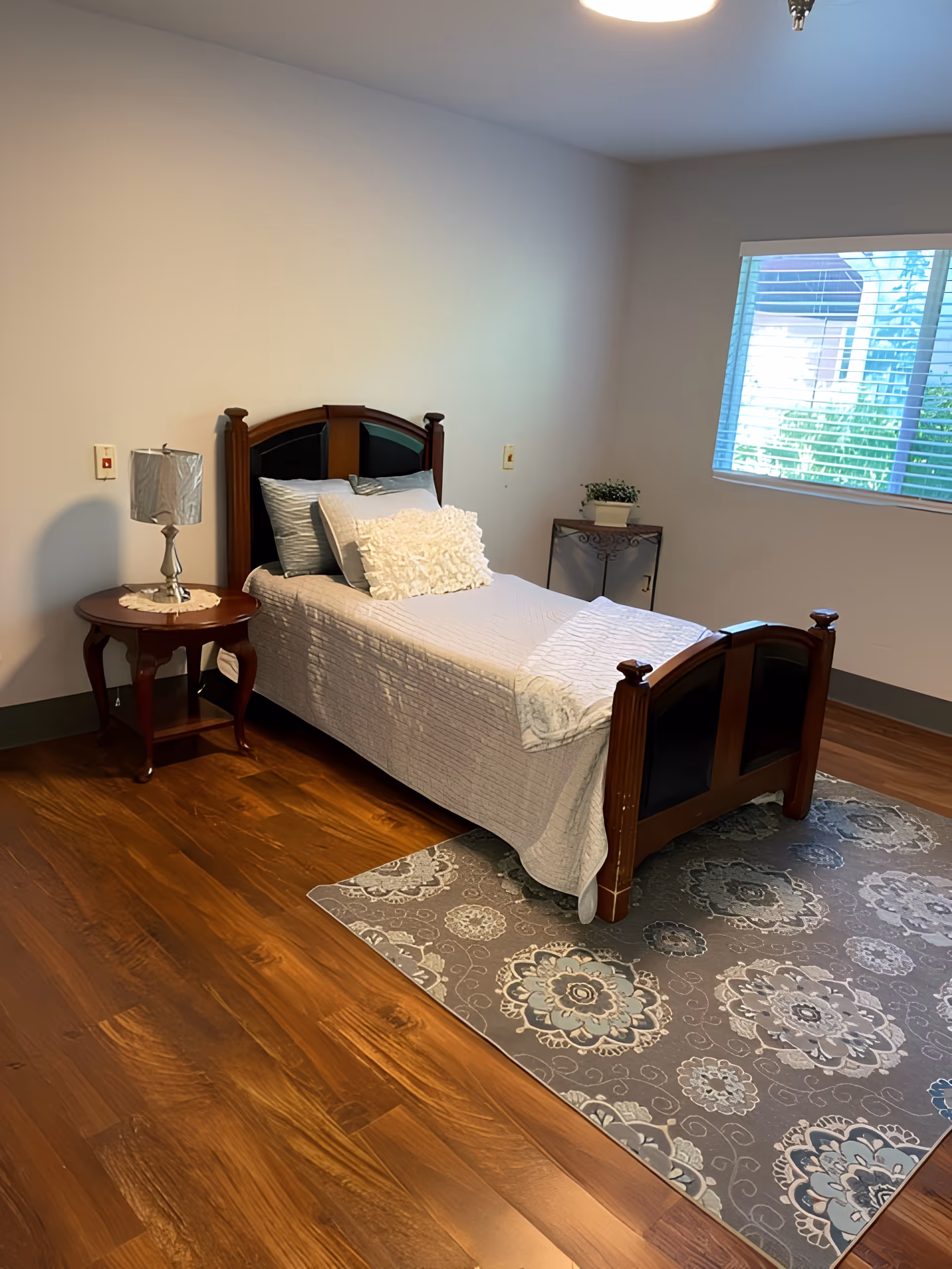 A tidy single bedroom with a wooden bed dressed in white bedding, a bedside table with a lamp, a patterned rug, and a window.