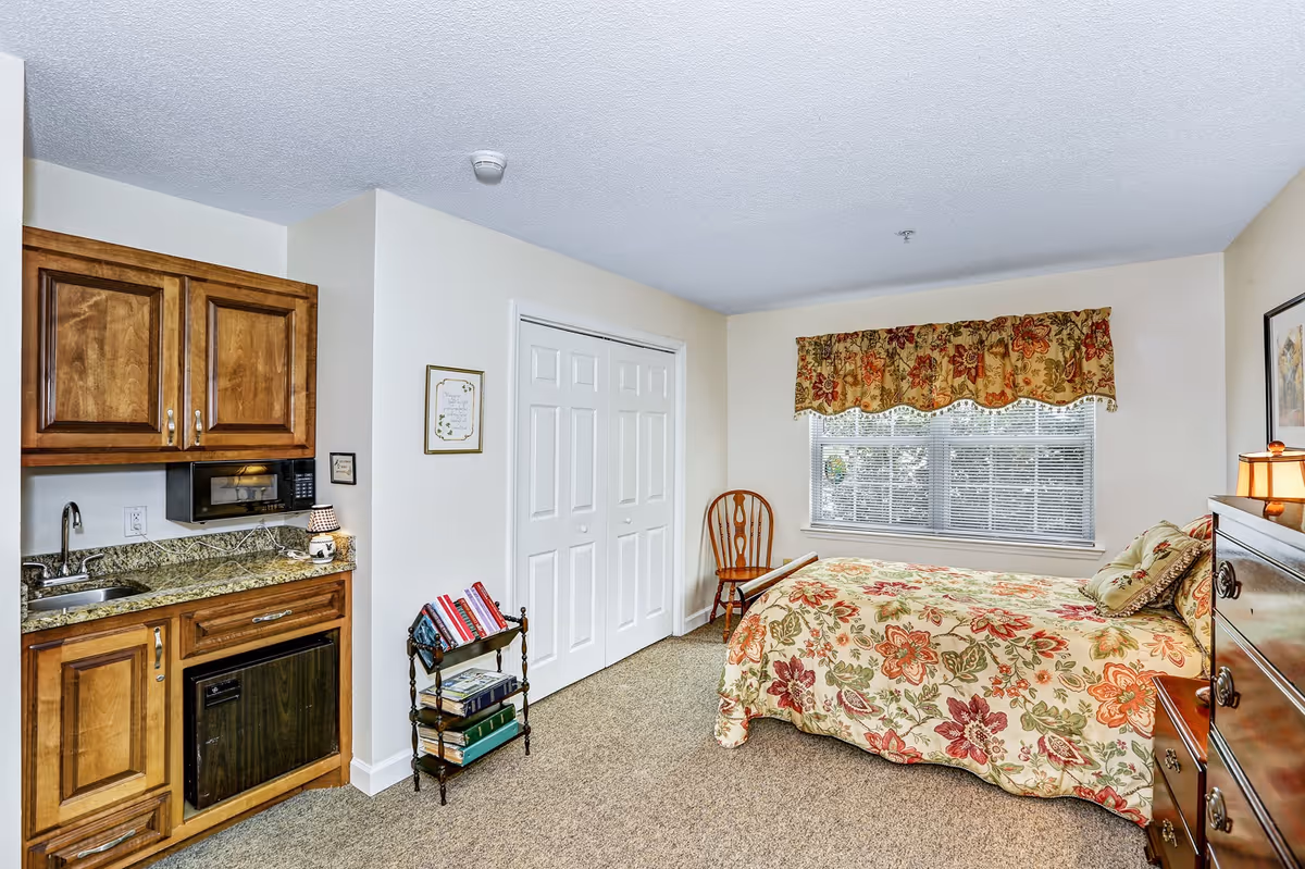 A cozy bedroom in a senior living facility featuring a bed with floral bedding and matching valance on the window. The room includes a wooden chair, a small bookshelf with books, a wooden dresser with a lamp, and a kitchenette area with wooden cabinets, a microwave, a small sink, and a mini fridge. The walls are light-colored and the floor is carpeted.