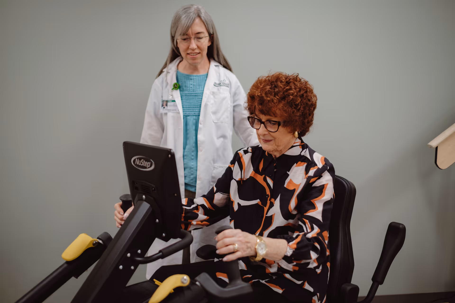 An elderly woman with curly hair and glasses is seated on a NuStep exercise machine, using the handles. A healthcare professional with long gray hair, glasses, and a white coat labeled Jordan Valley Senior Care stands behind her, observing and providing support in a clinical setting.