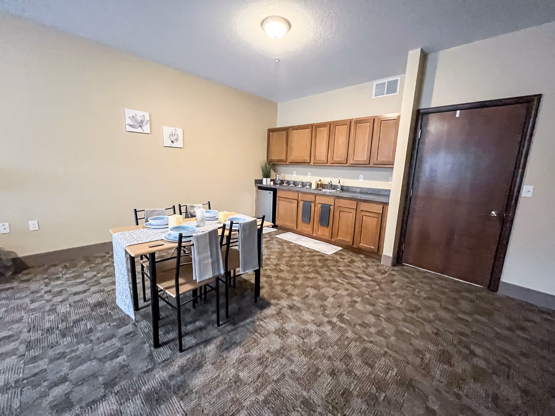 Interior view of a dining area in Reed City Fields featuring a small dining table set for four with plates, glasses, and candles. The room has carpeted flooring, beige walls with two floral paintings, wooden cabinets along one wall, a small refrigerator, and a closed wooden door.