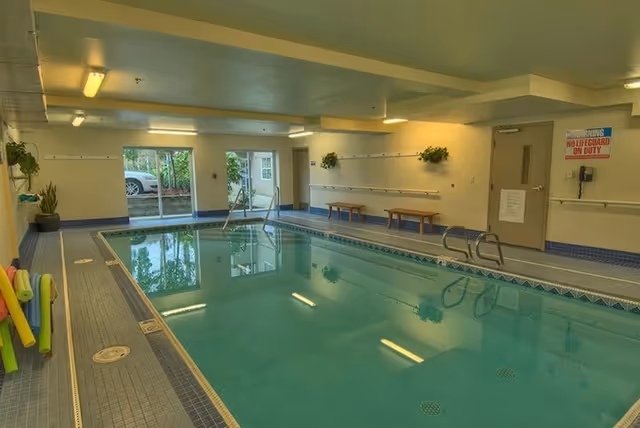 Indoor swimming pool room with benches, pool noodles, handrails and a 'No Lifeguard on Duty' sign.