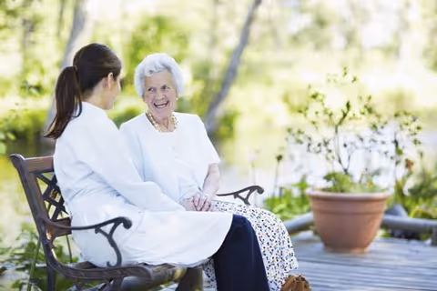 An elderly woman and a caregiver sitting on a wooden bench outdoors, engaged in a friendly conversation with greenery and a potted plant in the background.