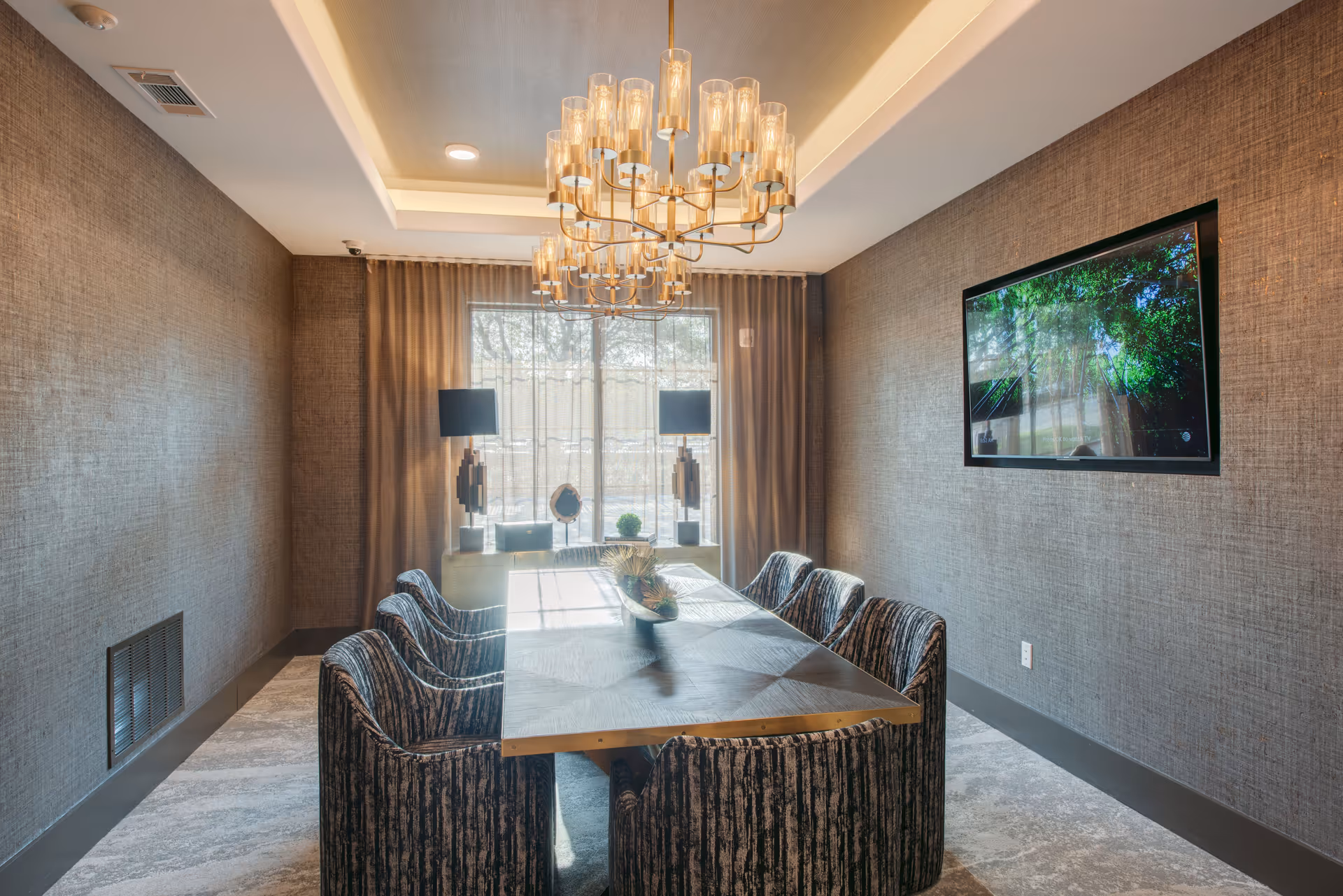A modern conference or dining room with a long rectangular table surrounded by eight upholstered chairs. The room features textured brown walls, a large window with sheer curtains, two decorative table lamps on a console beneath the window, and a flat-screen TV mounted on the wall. A stylish chandelier hangs from the ceiling above the table.