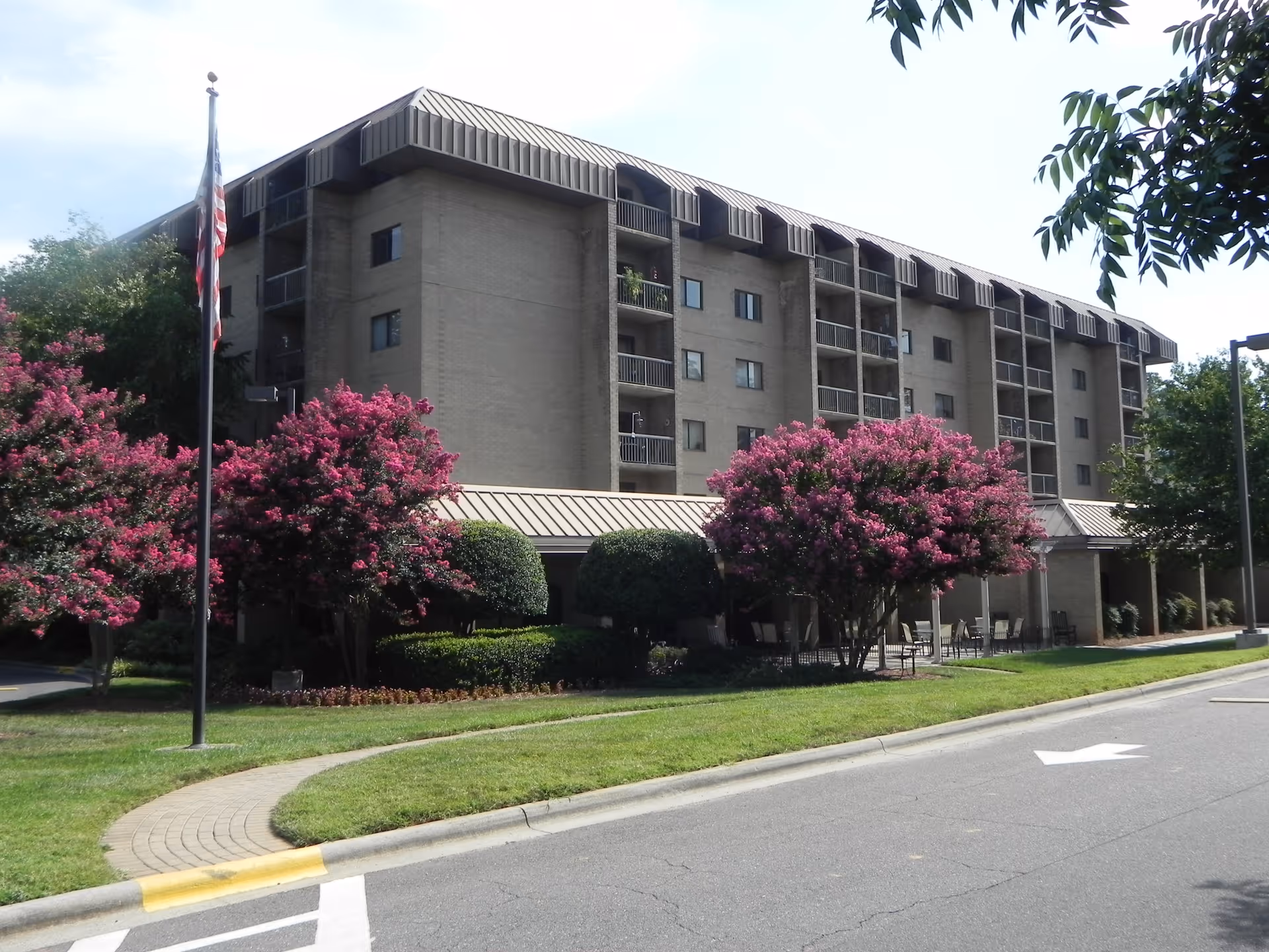 Exterior view of a multi-story senior living facility named Westminster Towers with balconies, surrounded by well-maintained landscaping including green bushes and trees with pink flowers, an American flag on a flagpole, and a paved driveway with a sidewalk.