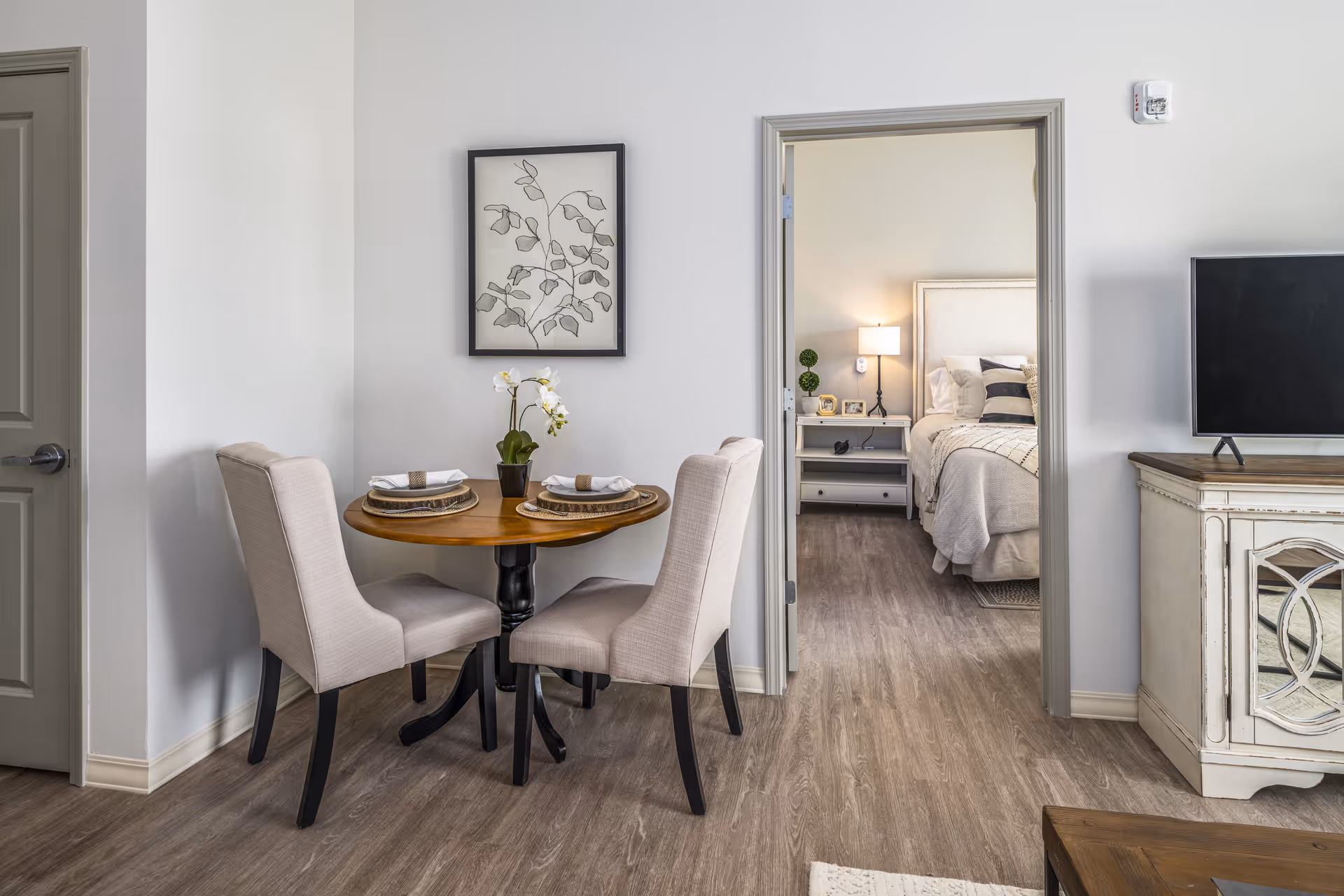 A small round dining table set for two with upholstered chairs in a bright apartment living area, a TV cabinet to the right and a doorway showing a furnished bedroom.