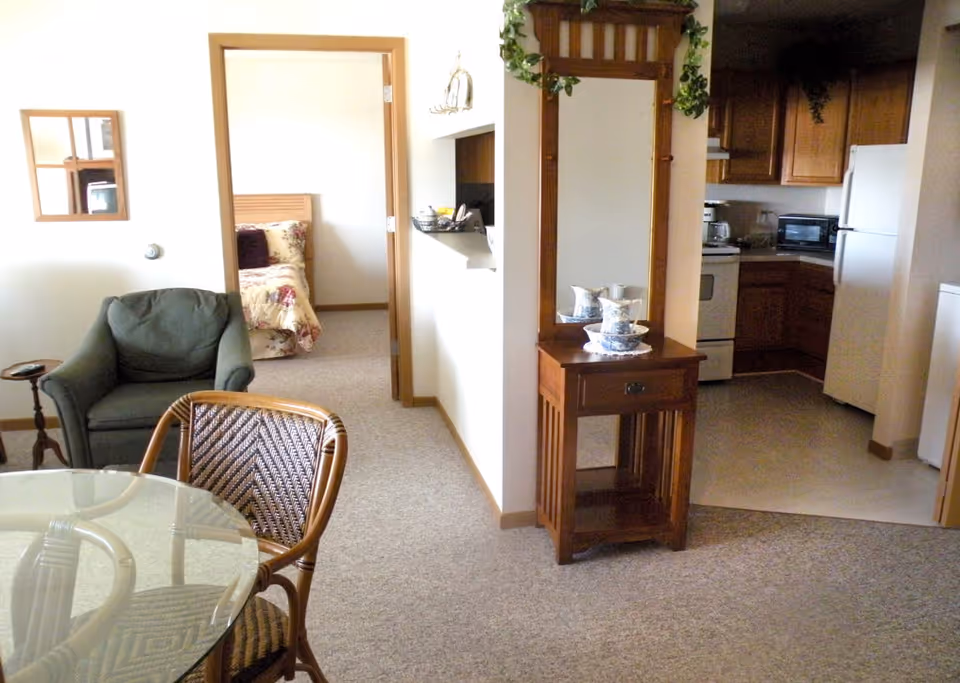 Interior view of a senior living apartment showing a small dining area with a glass-top table and wicker chairs, a green armchair, a wooden hall tree with a mirror and decorative items, a kitchen with wooden cabinets and white appliances, and a bedroom with a floral bedspread visible through an open doorway.