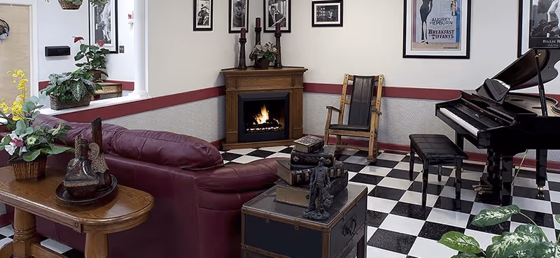 A cozy living room with a burgundy leather sofa, a wooden rocking chair, a black and white checkered floor, a fireplace with a fire burning, a grand piano with a bench, framed black and white photos and posters on the walls, and various plants and decorative items on tables.