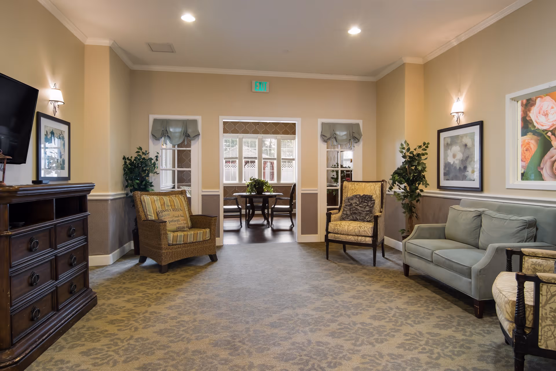 A cozy living room area in a senior living facility with a patterned carpet, a green sofa, two armchairs, framed floral artwork on the walls, potted plants, and a wooden cabinet with a mounted TV. In the background, there is a doorway leading to another room with a table and chairs near windows.