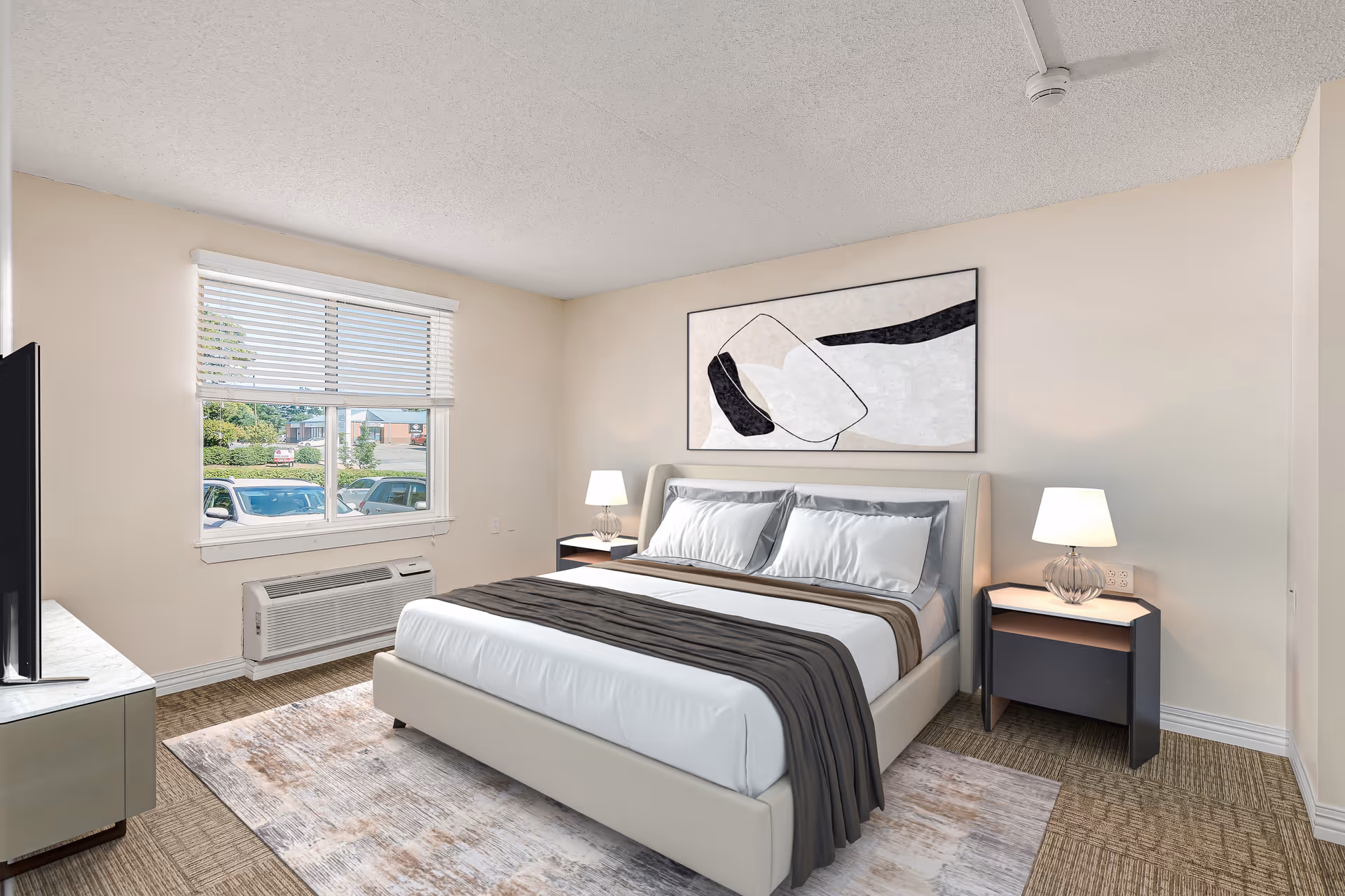 A bright bedroom with a large bed featuring white and gray bedding, two bedside tables each with a lamp, a large abstract painting above the bed, a window with blinds showing parked cars outside, and a TV on a stand opposite the bed.