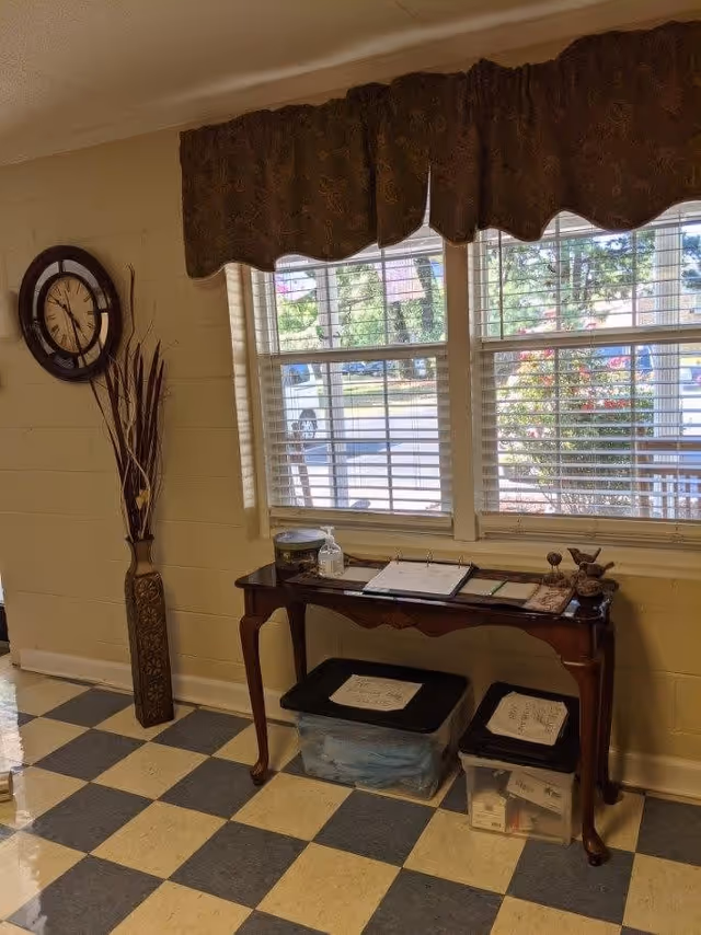 Interior view of a room with checkered floor tiles, a wooden table under a window with blinds and a brown valance. On the table are a hand sanitizer bottle, a container, and some papers. Below the table are two plastic storage bins with labels. A decorative tall vase with dried plants stands next to the wall, and a round wall clock is mounted above it.