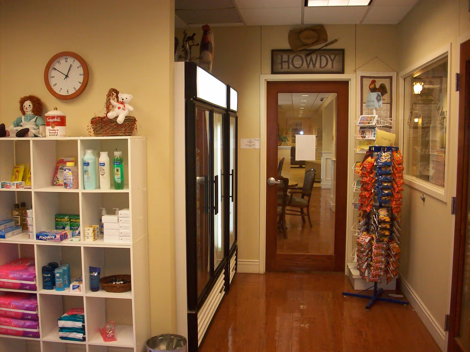 Interior hallway of a senior living facility with a white cubby shelf stocked with personal care items and snacks, a clock on the wall, a glass door with a 'Howdy' sign above it, and a rotating rack filled with snack bags.