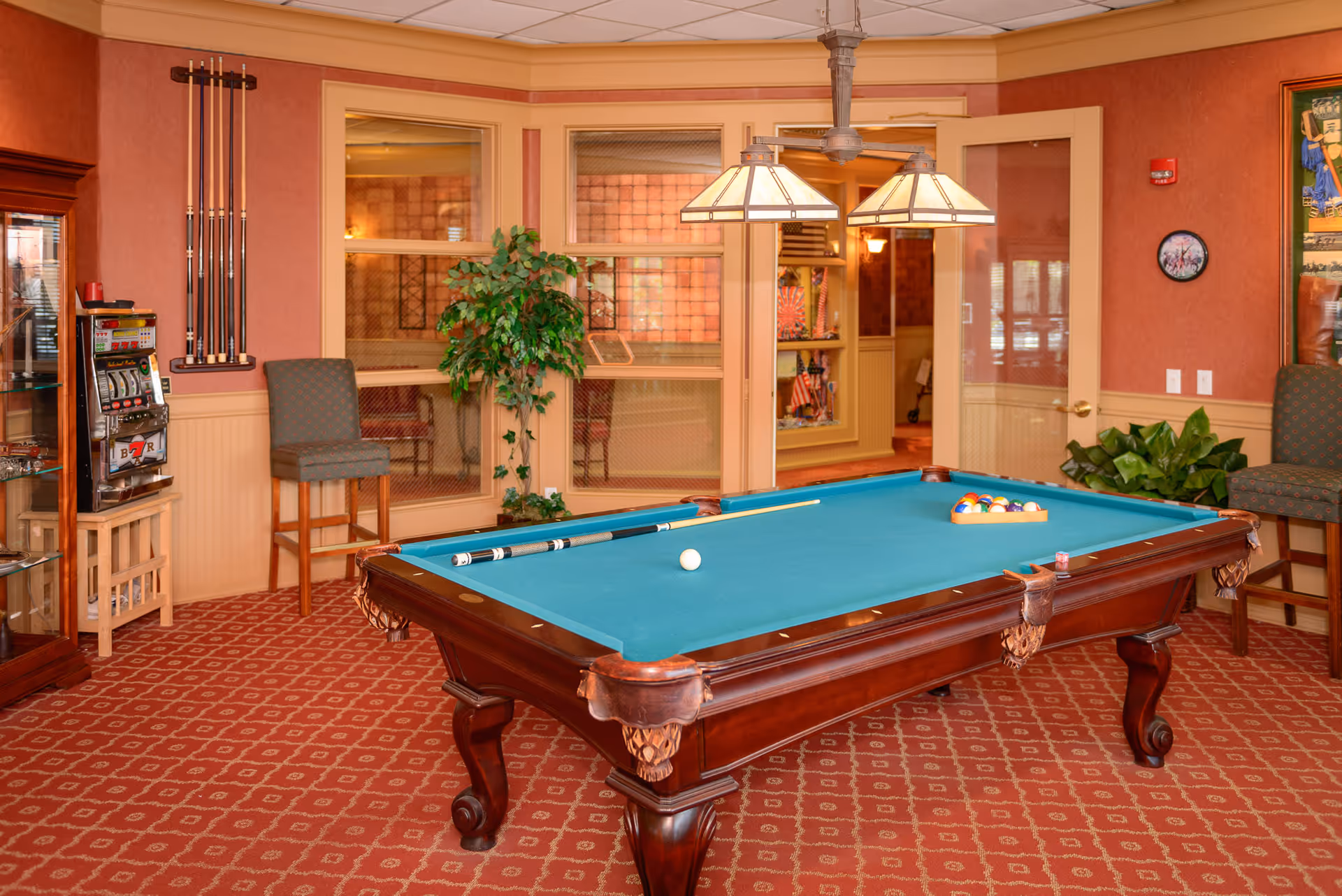 Carpeted game room interior with a pool table, cues, hanging lights, chairs, and plants.