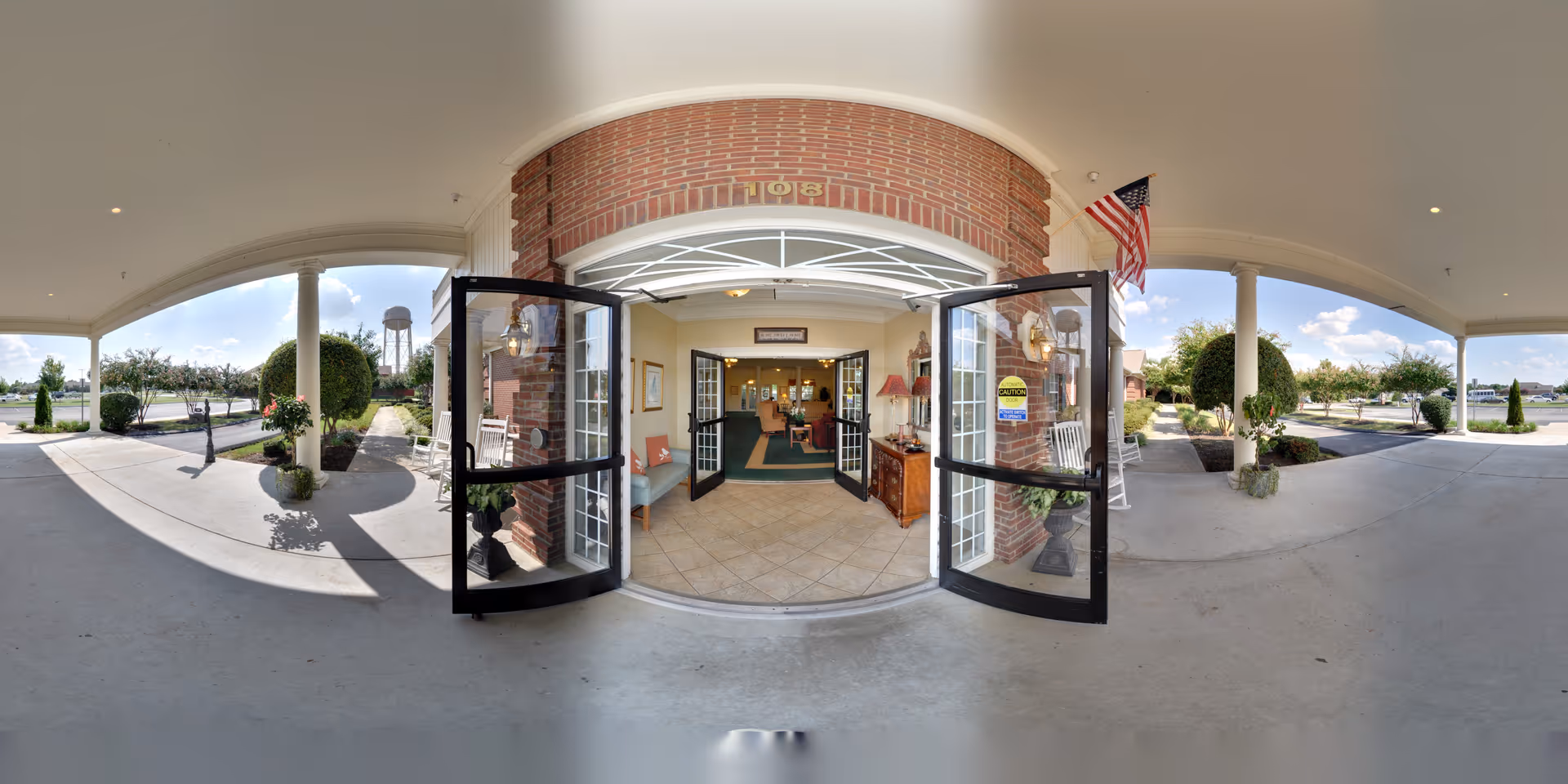 Entrance of The Pinnacle of Southaven facility showing double glass doors open under a covered porch with white columns. There are rocking chairs on either side of the entrance, neatly trimmed bushes, an American flag on the right, and a water tower visible in the background. Inside, a reception area with seating and lamps is visible.