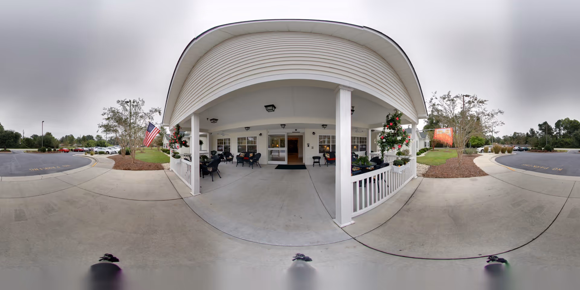 Wide-angle view of the covered entrance area of a senior living facility with white pillars and railing, several chairs and tables arranged for seating, hanging flower baskets, an American flag, and a parking lot with cars and trees in the background under a cloudy sky.