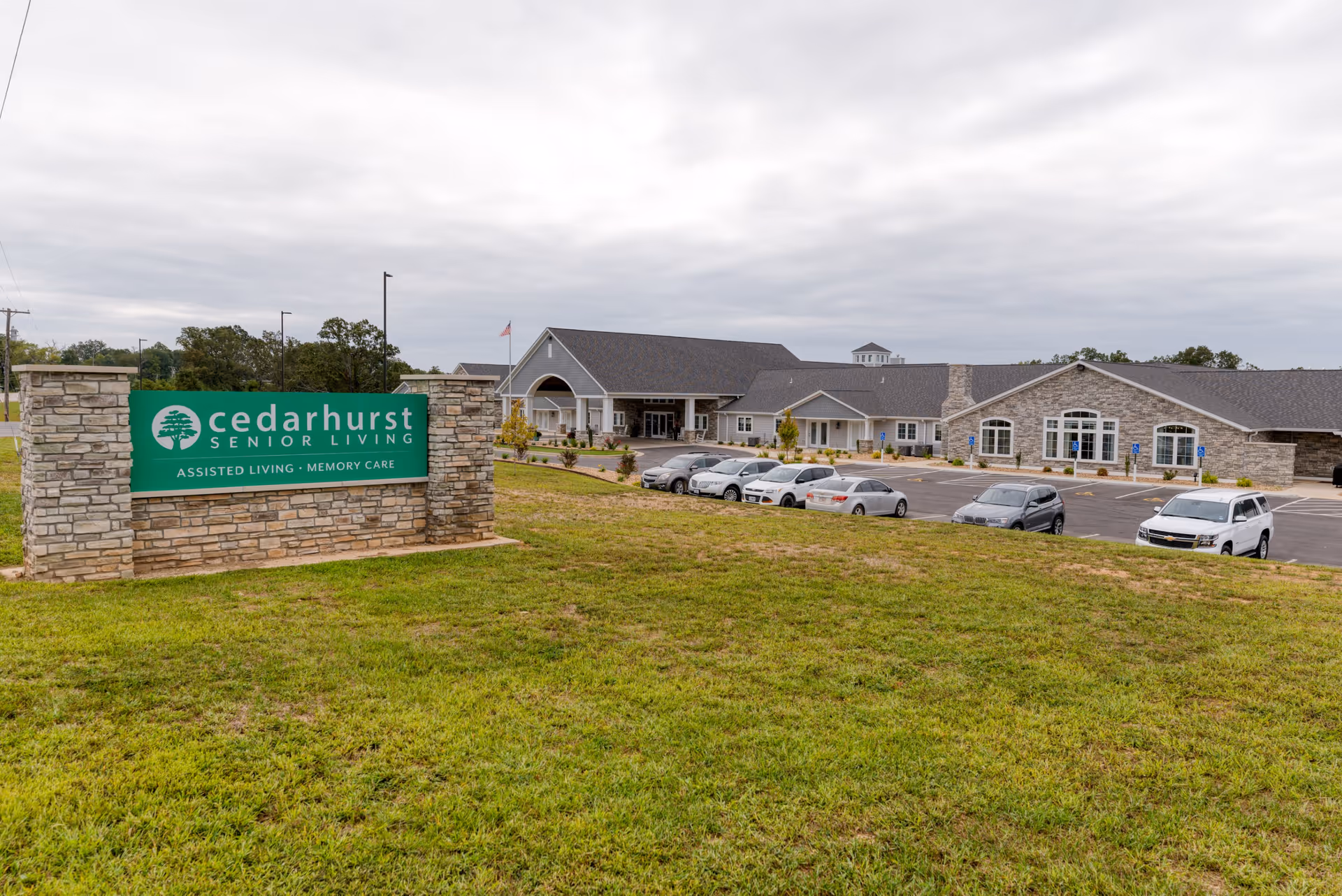 Exterior view of Cedarhurst Senior Living of West Plains facility with a large green sign in the foreground displaying the facility name and services. The building is a single-story structure with stone and siding, a covered entrance, and a parking lot with several cars parked. The sky is overcast.