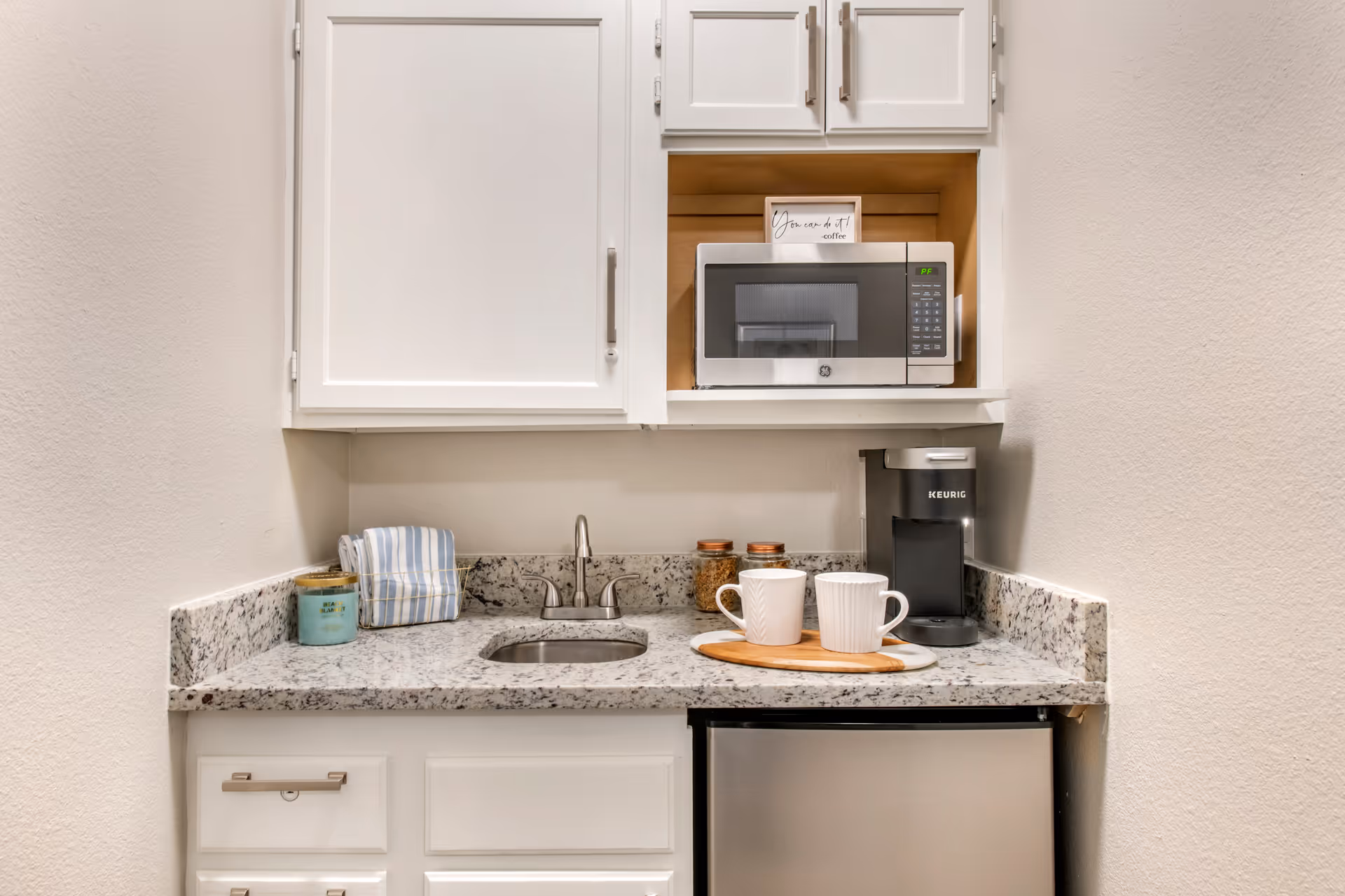 A small kitchenette area with white cabinets, a granite countertop, a stainless steel sink, a microwave, a Keurig coffee maker, two white mugs on a wooden tray, a small refrigerator, and a candle with a folded striped towel.