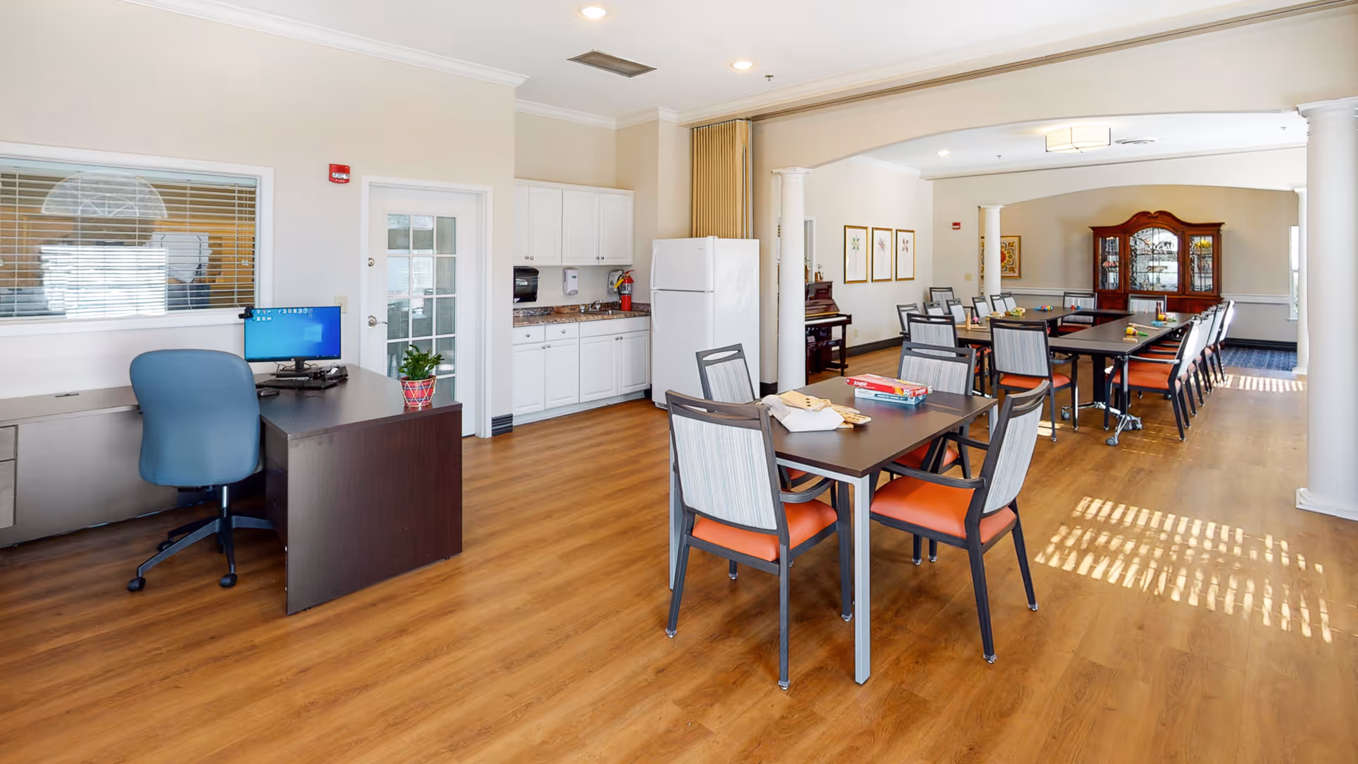 A spacious room with wooden flooring featuring a small kitchen area with white cabinets and a refrigerator, a dark wooden desk with a blue office chair and computer, and several tables with chairs arranged for group activities. The room has white walls, ceiling lights, and decorative columns, with framed artwork and a wooden cabinet in the background.