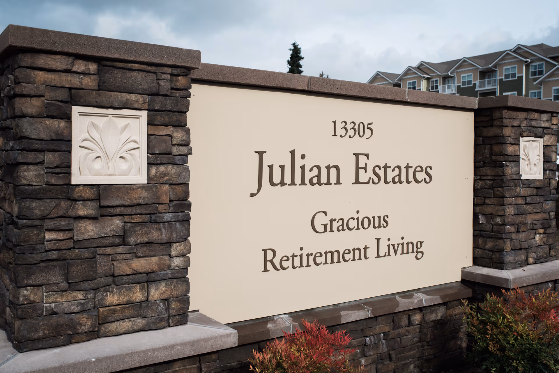Stone and concrete sign for Julian Estates Gracious Retirement Living with the address number 13305, set against a cloudy sky and some residential buildings in the background.