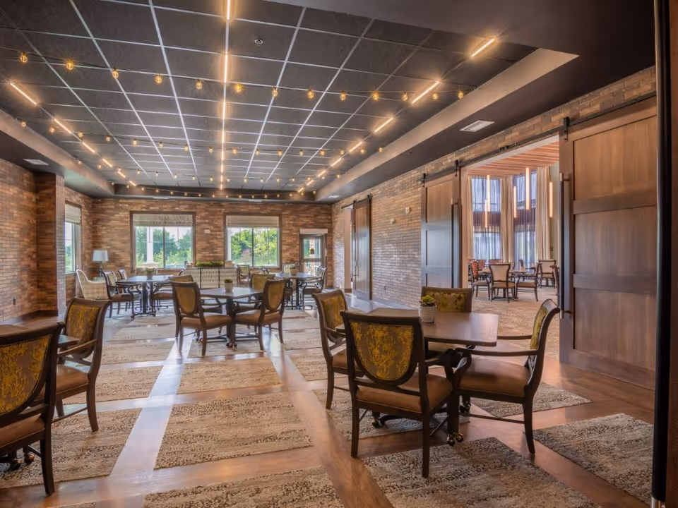 Spacious senior living dining room with multiple tables and chairs, brick walls, large windows, and string lighting.