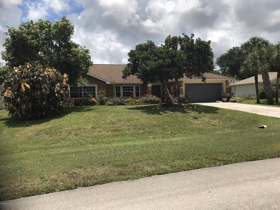 Single-story suburban house with a front lawn, trees, and an attached garage under a cloudy sky.