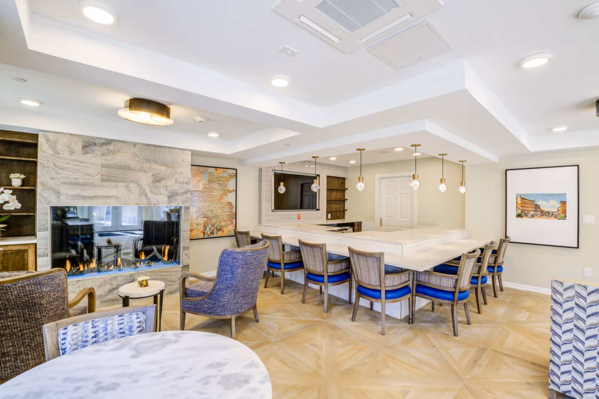 A bright and modern common area in a senior living facility featuring a large marble countertop bar with eight chairs that have blue cushions. There is a contemporary gas fireplace with a stone surround on the left wall, flanked by built-in shelves. The room has wood-patterned flooring, recessed lighting, and pendant lights hanging above the bar. Artwork is displayed on the walls, and there are additional seating areas with upholstered chairs and a round marble table in the foreground.