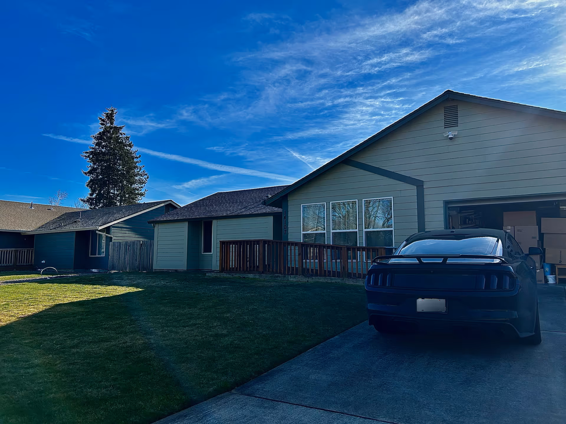 Front exterior of a single-story house with a lawn, driveway, and a car parked by an open garage under a blue sky.