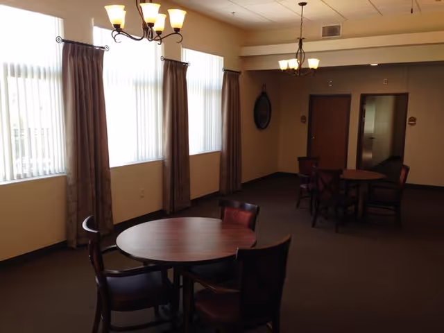 Interior view of a common area with round wooden tables and chairs, beige walls, large windows with vertical blinds and curtains, and ceiling light fixtures.