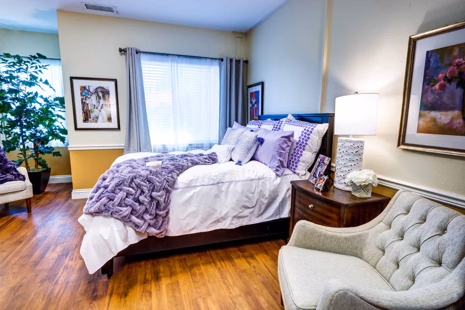 A cozy, well-lit bedroom with a bed dressed in white linens and purple accents, a nightstand with a lamp, and an upholstered armchair.
