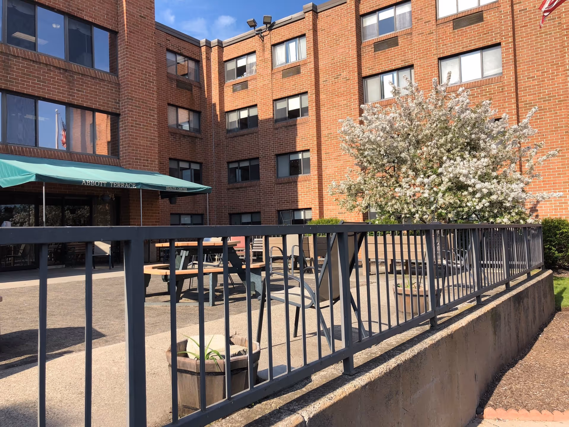 Outdoor patio area of Abbott Terrace Health Center with metal railing in the foreground, wooden benches, chairs, a flowering tree, and a brick building with multiple windows in the background under a clear blue sky.